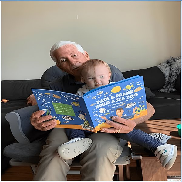 An elderly man reading Paul & Frank Build a Sea Zoo by Thomas Rippy to a toddler while sitting on a chair in a cozy living room.
