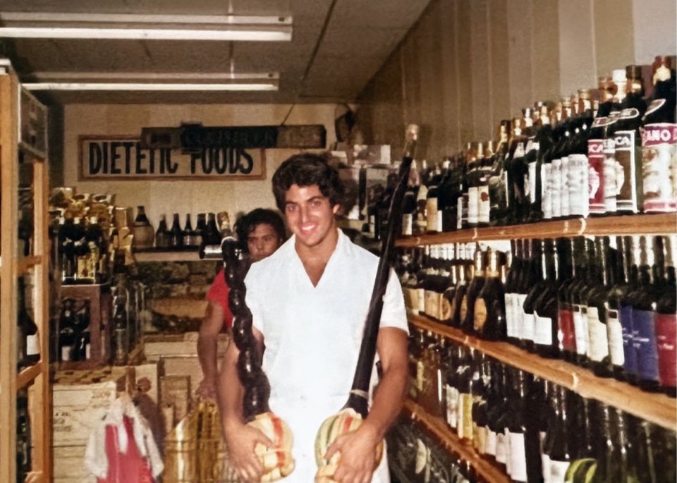 Thomas Rippy smiling and posing with a decorative item inside a vintage dietetic foods and wine store, surrounded by shelves of wine bottles.