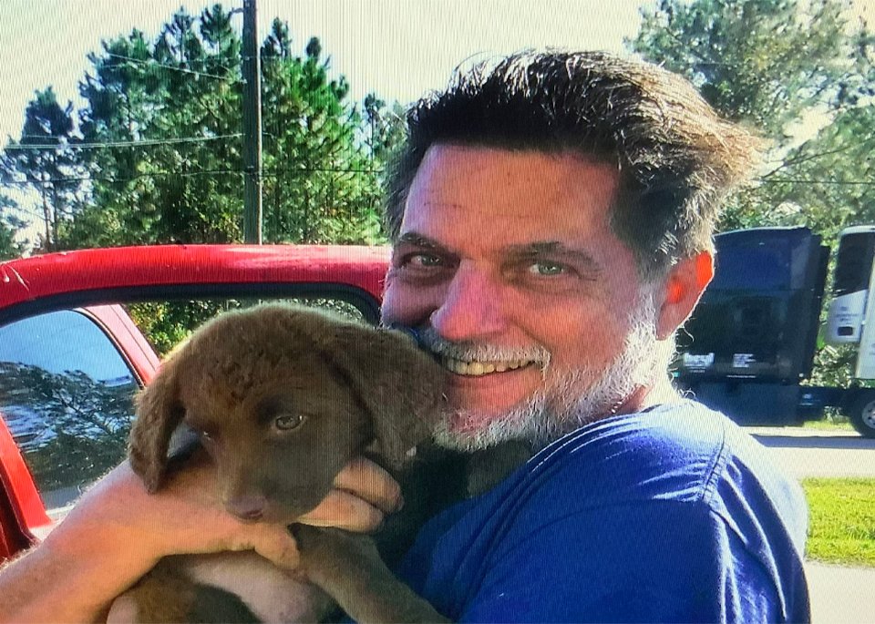 Thomas Rippy smiling warmly while holding a brown puppy outdoors next to a red vehicle.